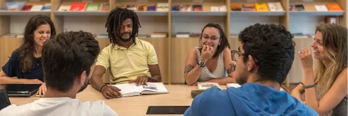 Six young adults sitting round a desk chatting, smiling and laughing. In the background are shelves of books